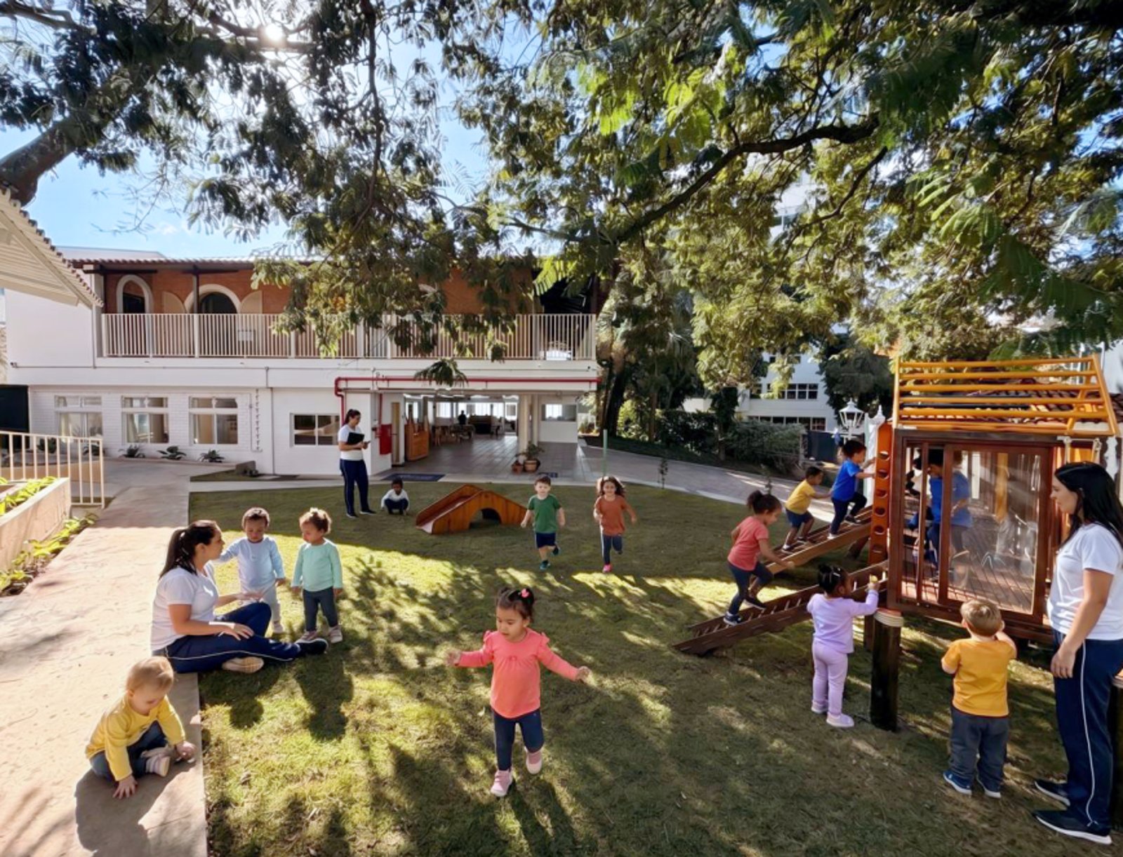 Crianças e educadoras no quintal da escola, com gramado, árvores e a fachada ao fundo