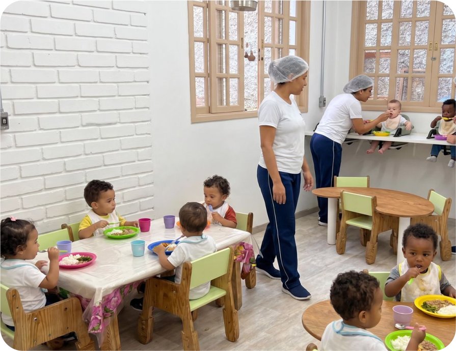 Crianças pequenas sentadas à mesa durante a refeição, acompanhadas por educadoras