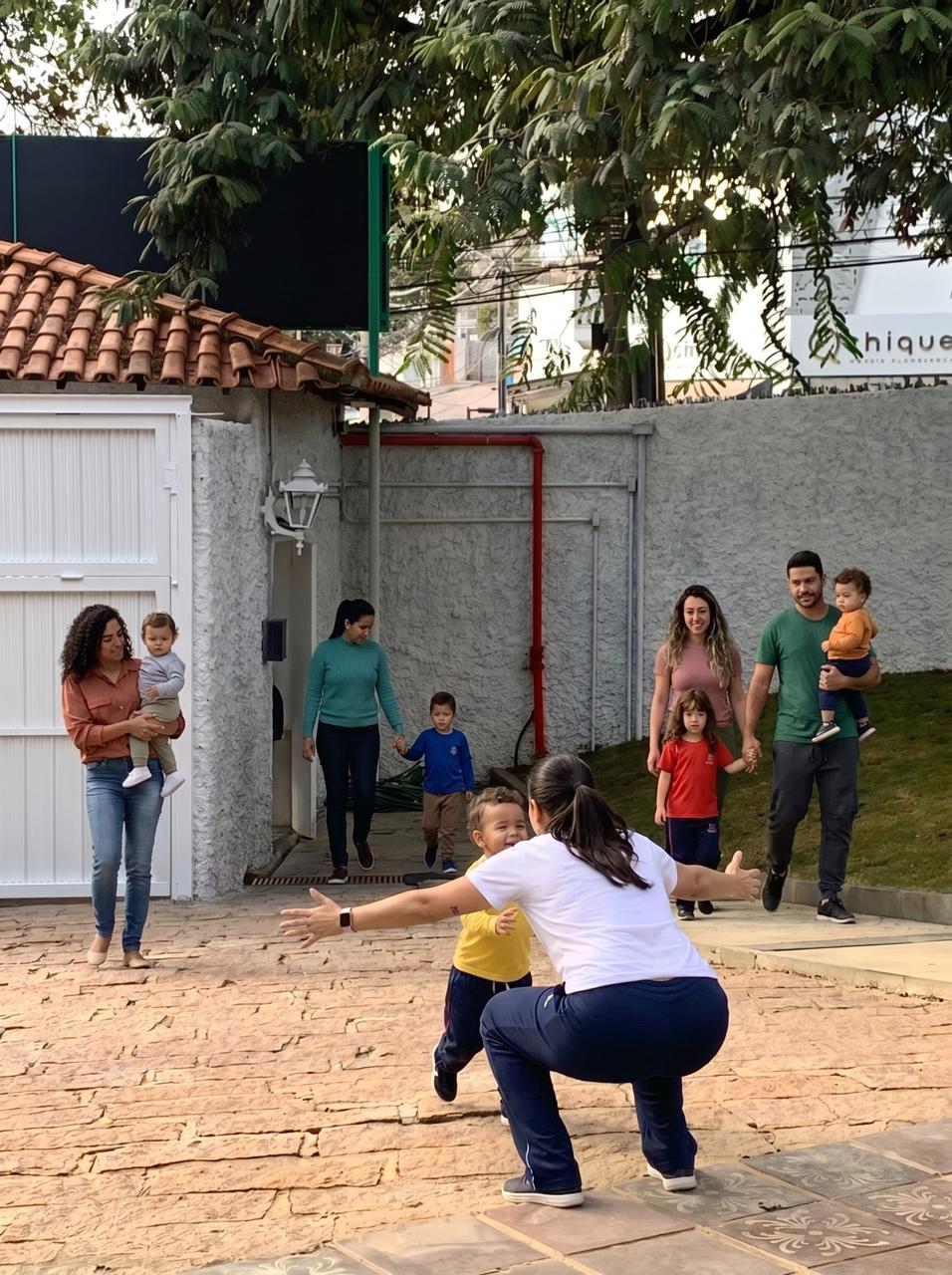 Educadora acolhendo uma criança na entrada da escola, com famílias ao fundo